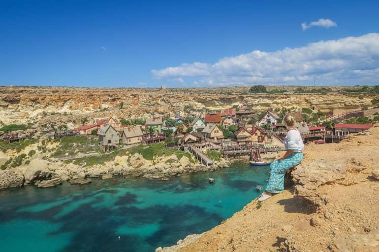 Woman sits on a rocky ledge looking down at Popeye Village - a film set with colorful wooden houses
