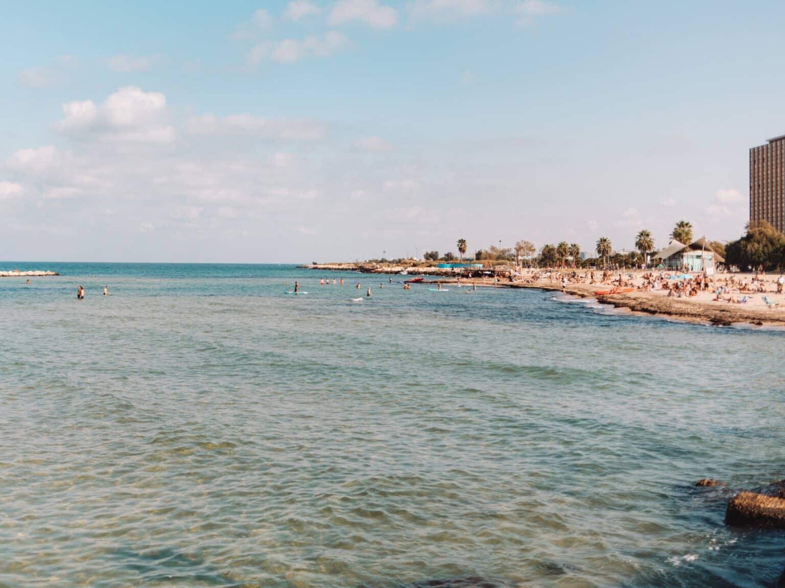 Turquoise water at Spiaggia Pane e Pomodoro beach, with palm trees and buildings in the background.