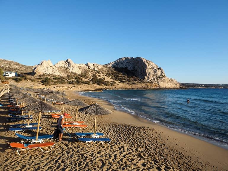 At Karpathos beach Ágios Theódoros, straw umbrellas and blue and orange sun loungers line the sandy beach, with a rock formation rising out of the dark blue sea in the background.
