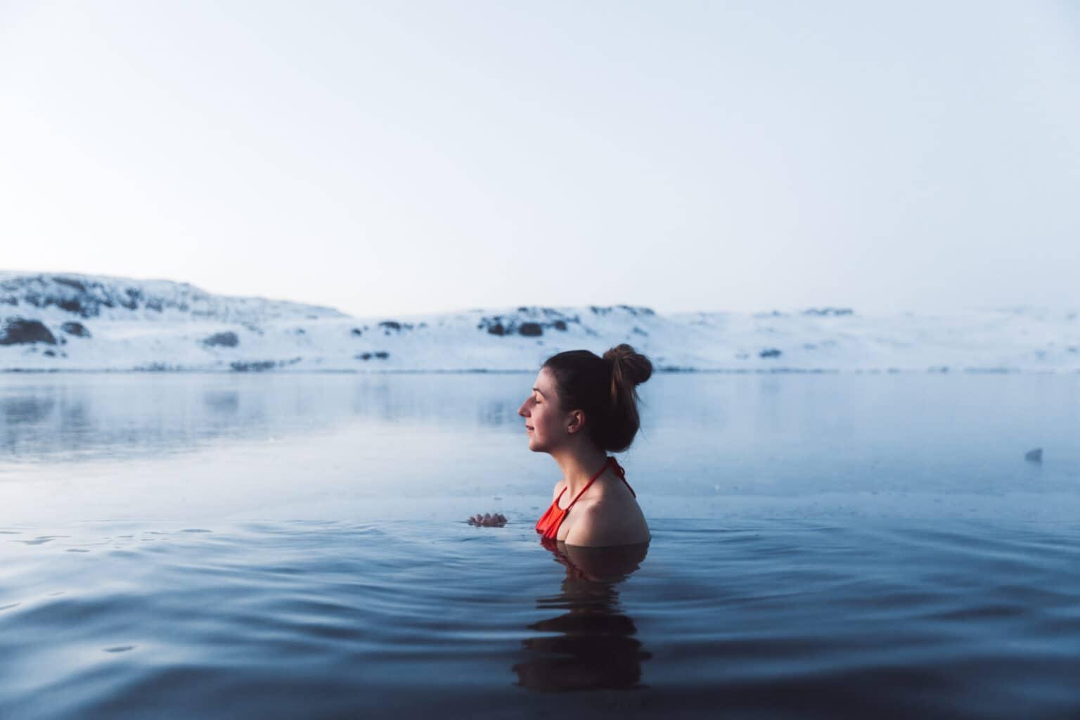 Young woman in a red swimsuit bathing in a hot thermal bath with a view of the snow-covered mountain peaks in North Iceland.