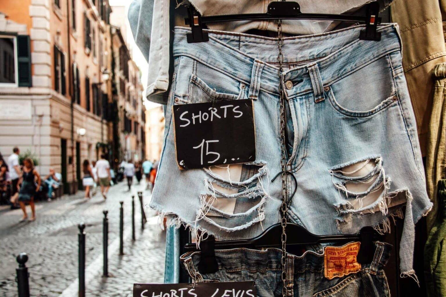 En primer plano cuelga un pantalón corto de mezclilla con etiqueta de precio, al fondo la gente pasea por las calles del barrio Monti en Roma.