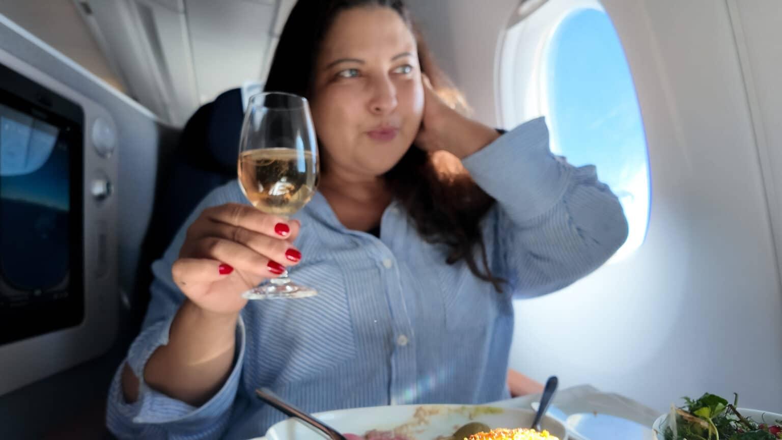 A woman sits in a Business Suite on the airplane. She holds a glass of champagne in her hand, and a warm meal is served in front of her.