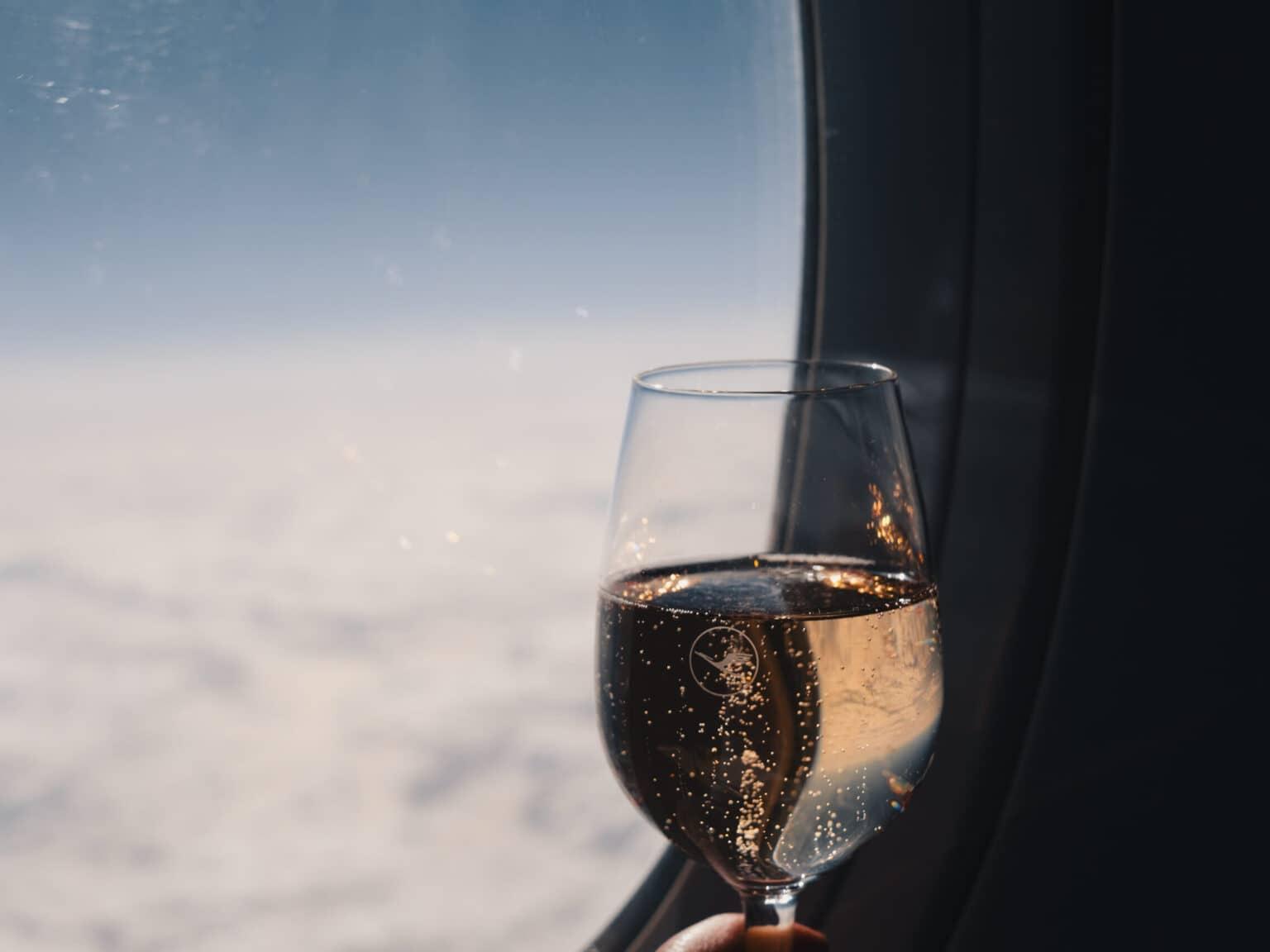A glass of champagne in front of an airplane window. In the background, there's a white cloud cover and above it, a blue sky.