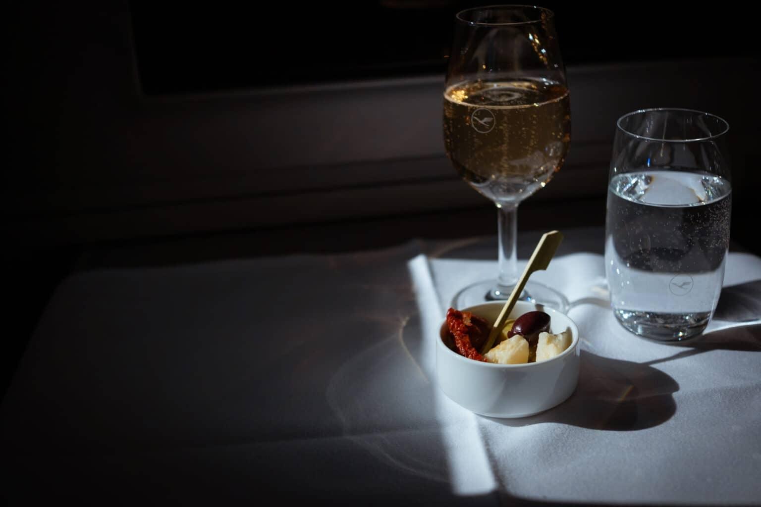 On a small table with a white tablecloth is a glass of champagne, a glass of water, and a small bowl with cheese, olives, and sun-dried tomatoes.