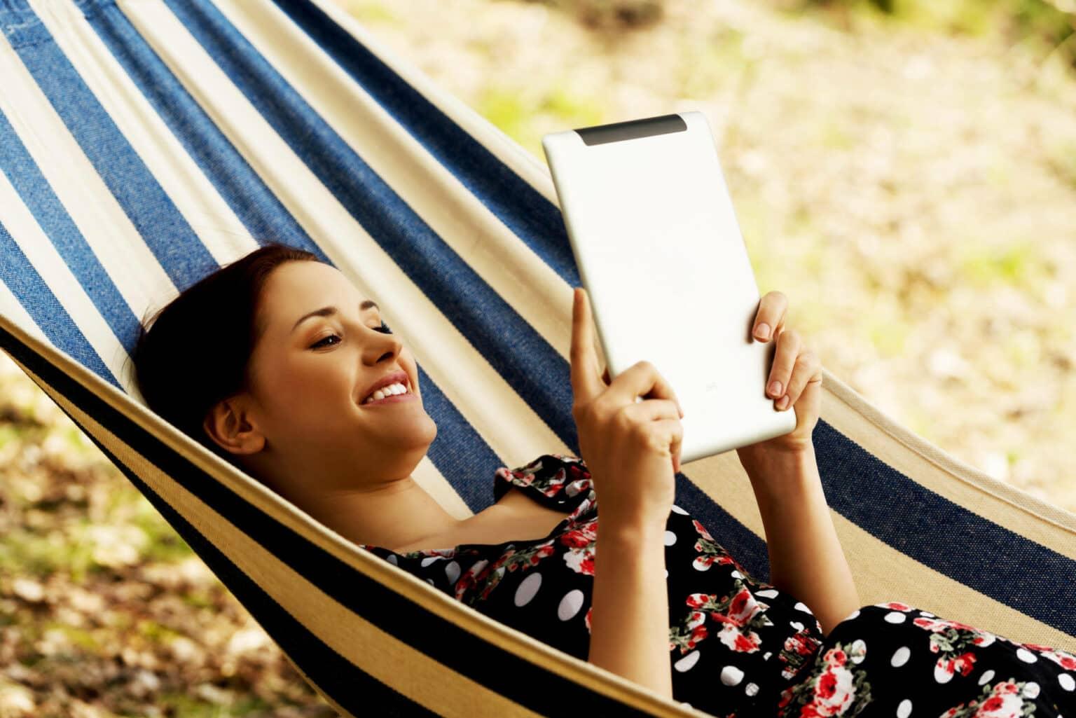 A woman smiles while reading her eBook from the hammock.