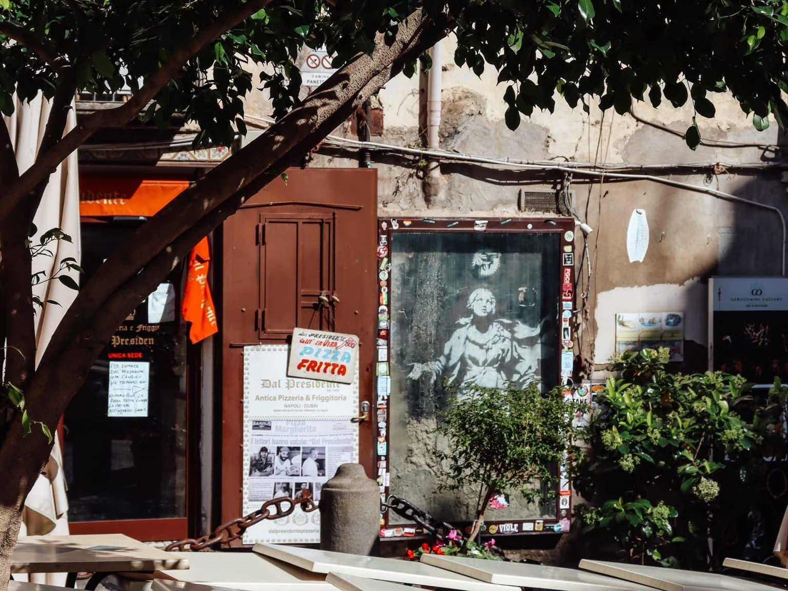 In Banksy's artwork, we see Madonna with a gun, with café tables and a tree in the foreground.