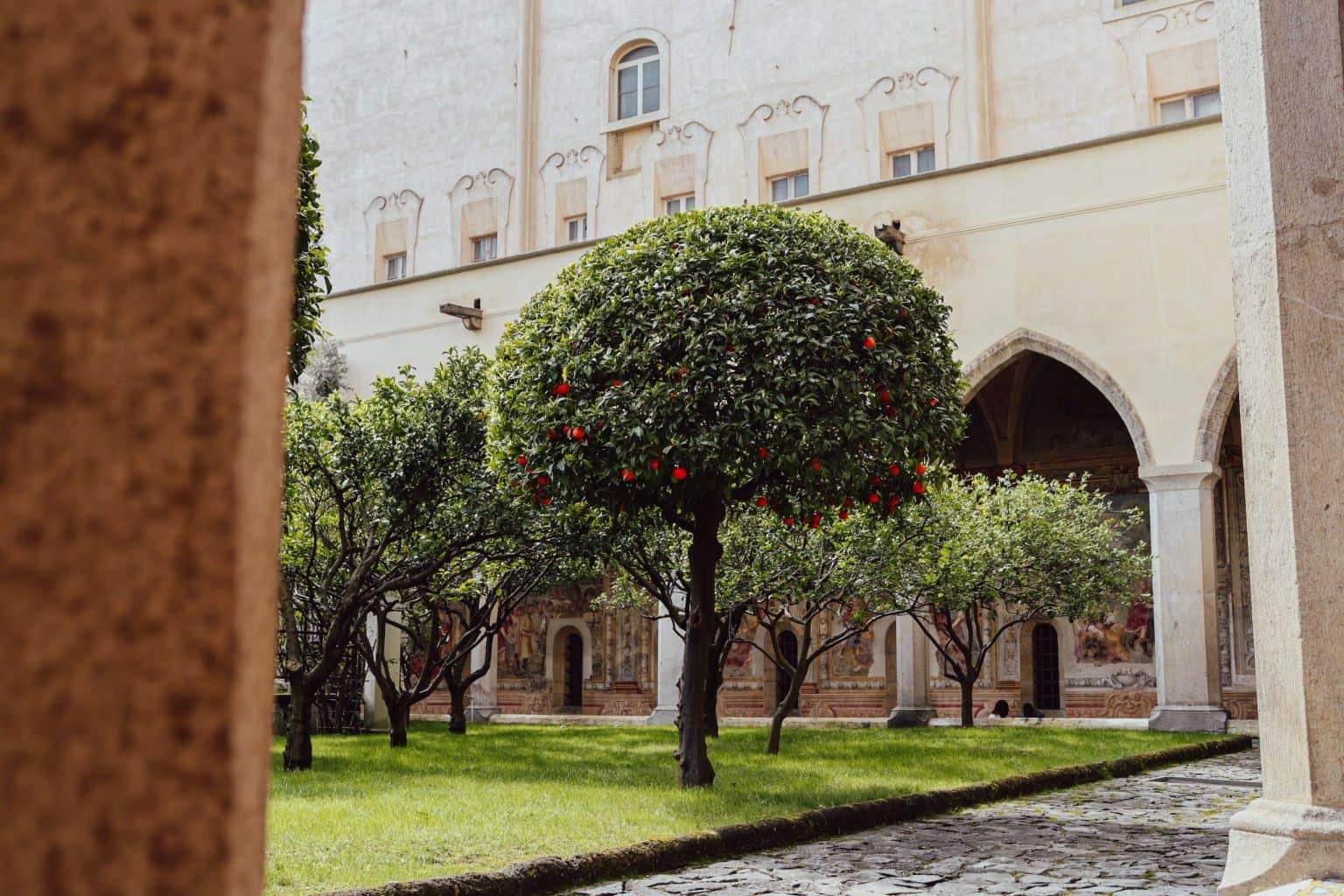 A sandstone-colored cloister with Gothic arches in the garden of Santa Chiara, with small trees on a lawn in front.