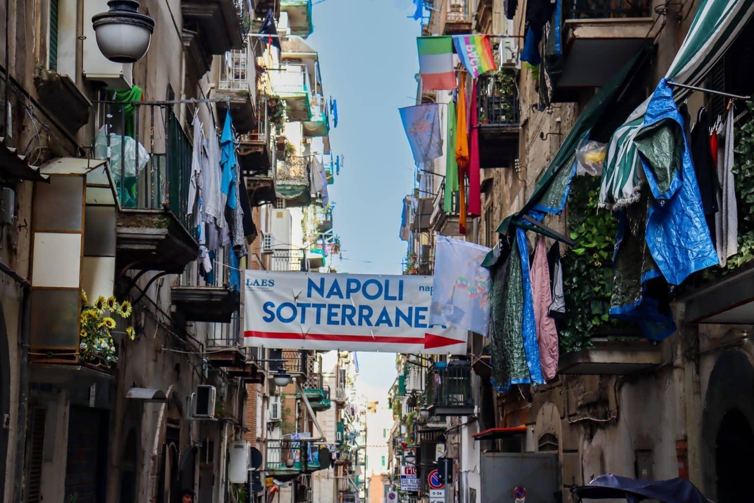 A narrow alley adorned with numerous balconies filled with laundry and flags, including one promoting 'Napoli Sotterranea'.