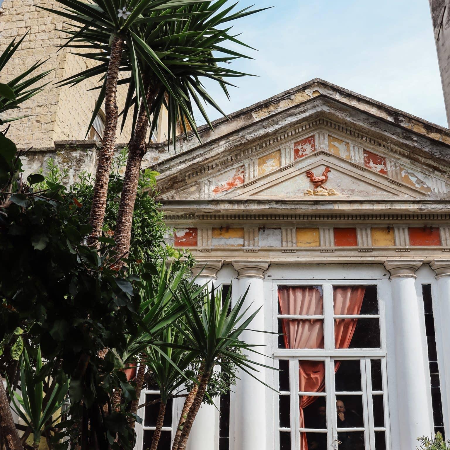 A windowed doorway of Palazzo Venezia adorned with columns, in front of which are palm trees.