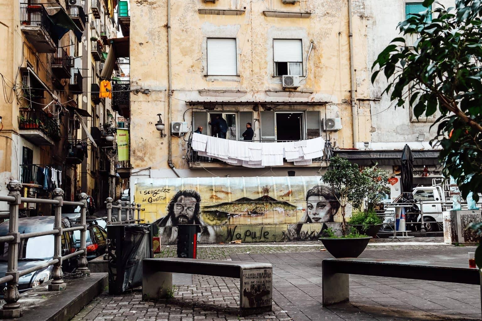 A dilapidated-looking Neapolitan building facade with graffiti, with two benches in the foreground.