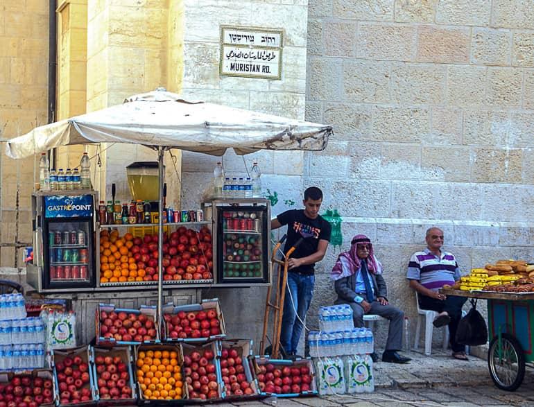 En una esquina de Jerusalén, Israel, y bajo un paraguas blanco, un comerciante ha montado un puesto en la calle con frutas en cajas para la venta, dos hombres mayores en sillas de plástico esperan clientela.