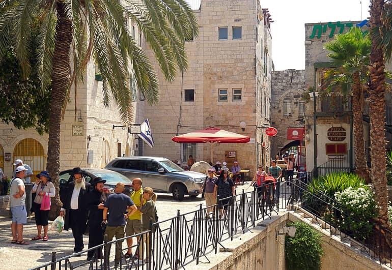 En el barrio judío de Jerusalén, transeúntes caminan por las fachadas de casas de color piedra arenisca y palmeras más allá.