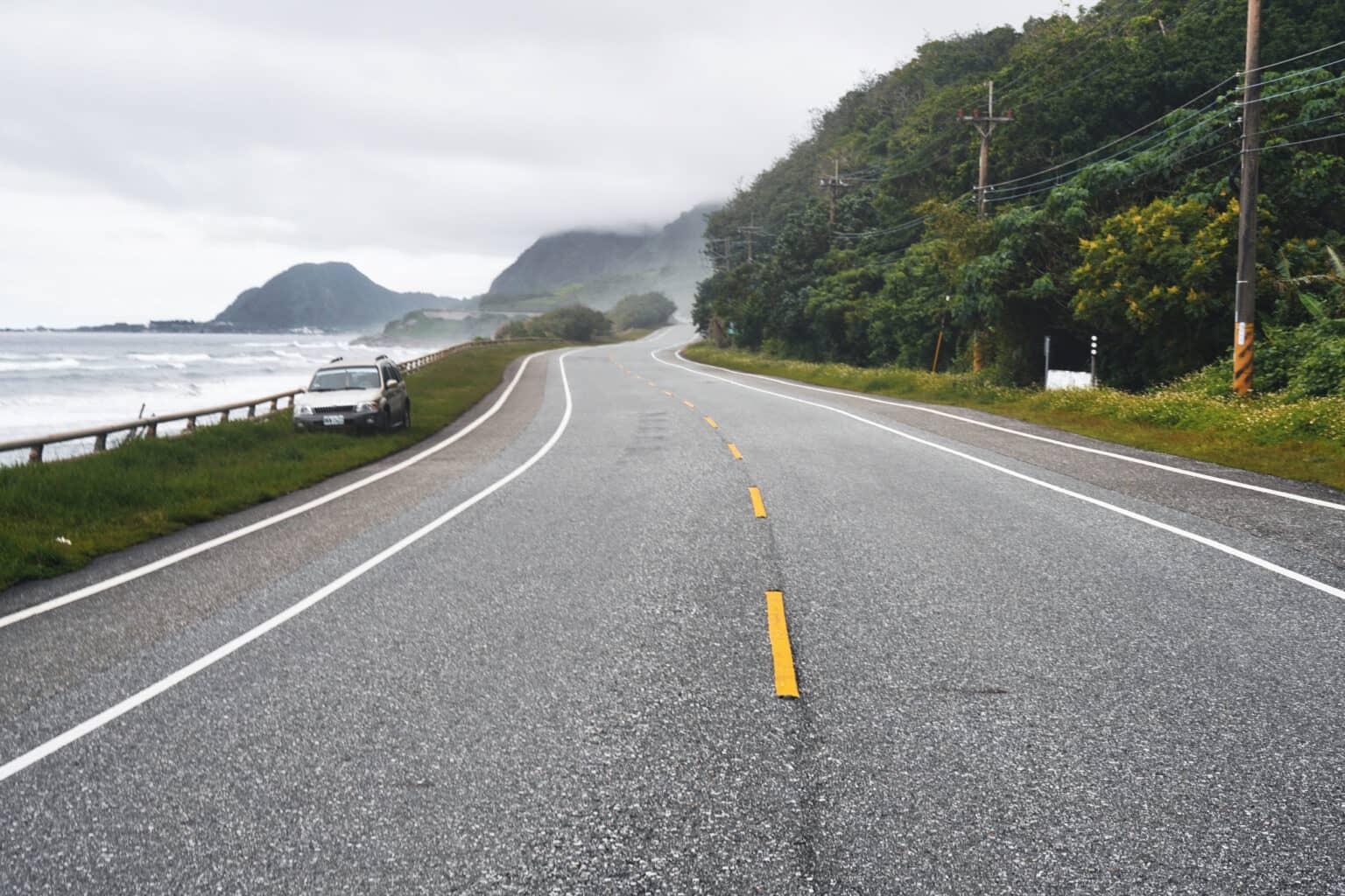A street right by the sea in Taiwan with a silver car parked by the roadside.
