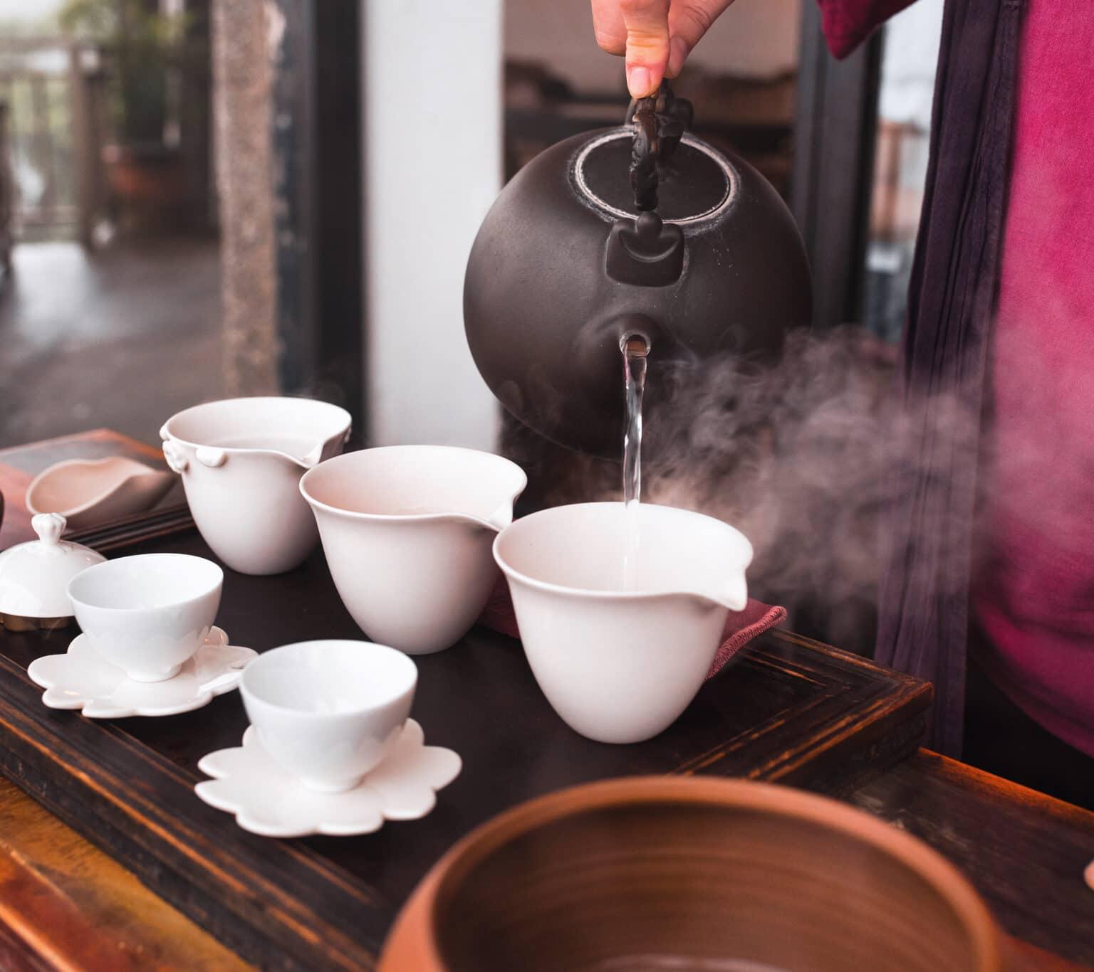Boiling water is poured from a black teapot into white cups.