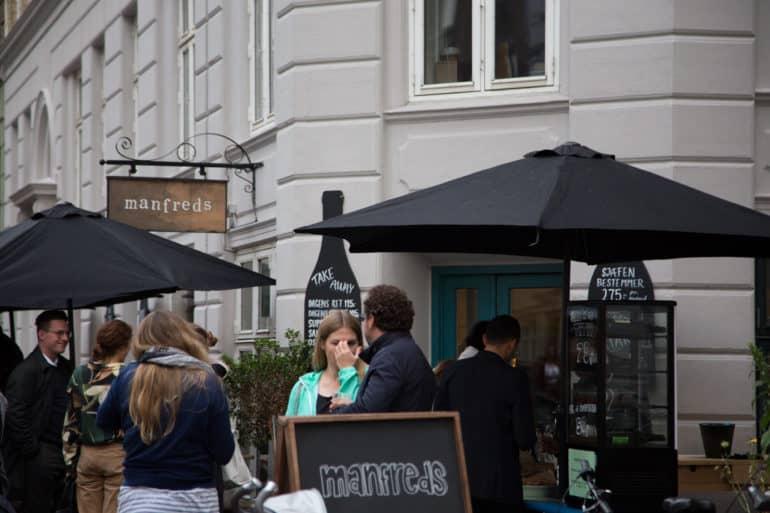 Frente a la entrada del bar Manfreds en Copenhague, hay personas bajo sombrillas negras charlando.