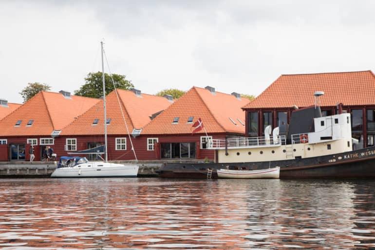 Un velero y un barco están anclados frente a una costa en Copenhague, al fondo se ven casitas rojas con ventanas blancas.