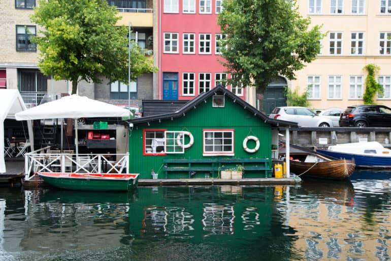 En un canal de Copenhague hay un barco familiar verde con una pequeña terraza y una sombrilla blanca, en la fachada de la casa cuelgan dos salvavidas.