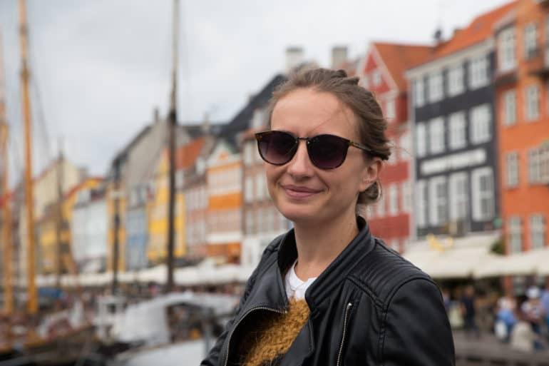 La bloggera Anika sonríe con su pelo castaño y unas gafas de sol negras y una chaqueta de cuero ante la cámara, en el fondo se vislumbra borroso la fachada de los edificios del centro de Copenhague.