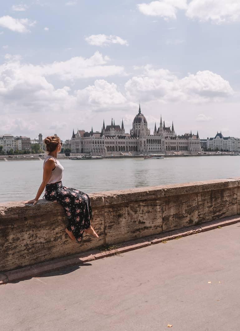 Viaje a Budapest: Una mujer sentada en un muro, al fondo se ve agua y un edificio.