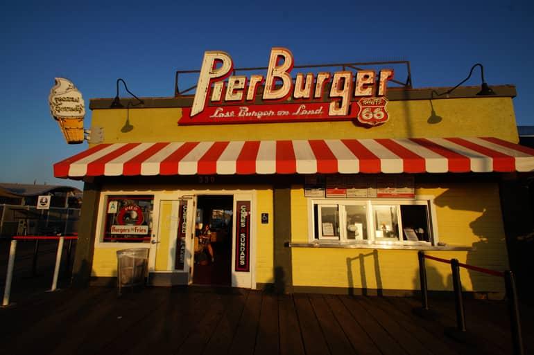 A yellow burger shack with the sign Pier Burger and a red-and-white striped awning at sunset in Santa Monica.