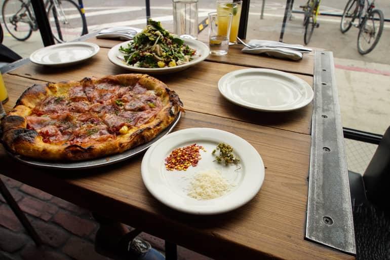 In Santa Monica, at Venice Beach, the restaurant Gjelina serves wood-fired pizza, with a plate of spices and a salad provided on the table.