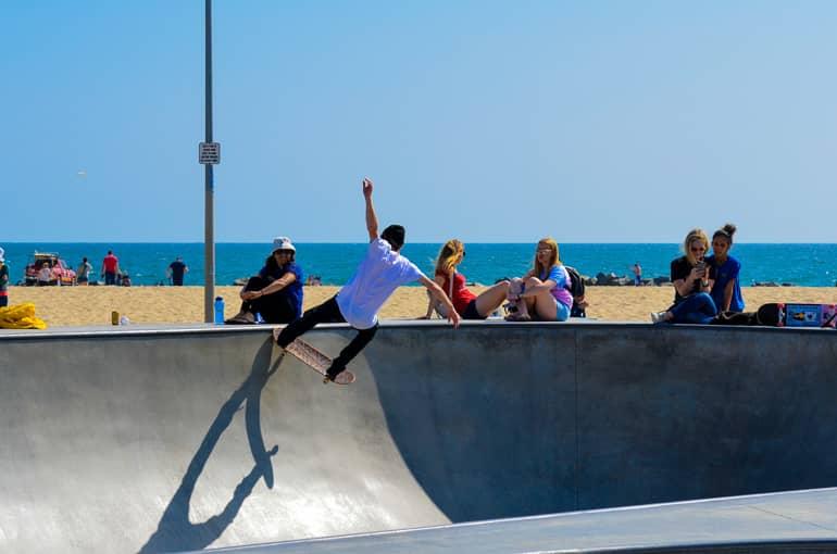 A skater on a skate bowl at Venice Beach in Santa Monica showcases his skills to a few interested spectators, with the beach and sea as his backdrop.