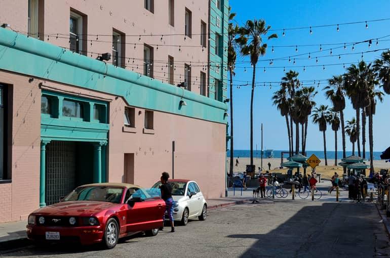 A side street in Santa Monica at Venice Beach shows a pink and green house facade, with a woman getting into her red Ford Mustang, string lights and palm trees reaching into the sky.