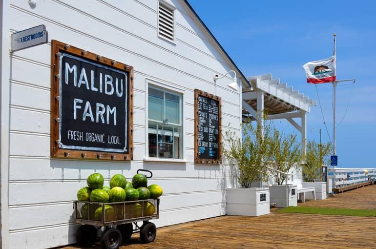 The Malibu Farm restaurant with its whitewashed facade is located on the Malibu Pier, with a small cart filled with watermelons next to its wall, and a Los Angeles flag waving in the wind.