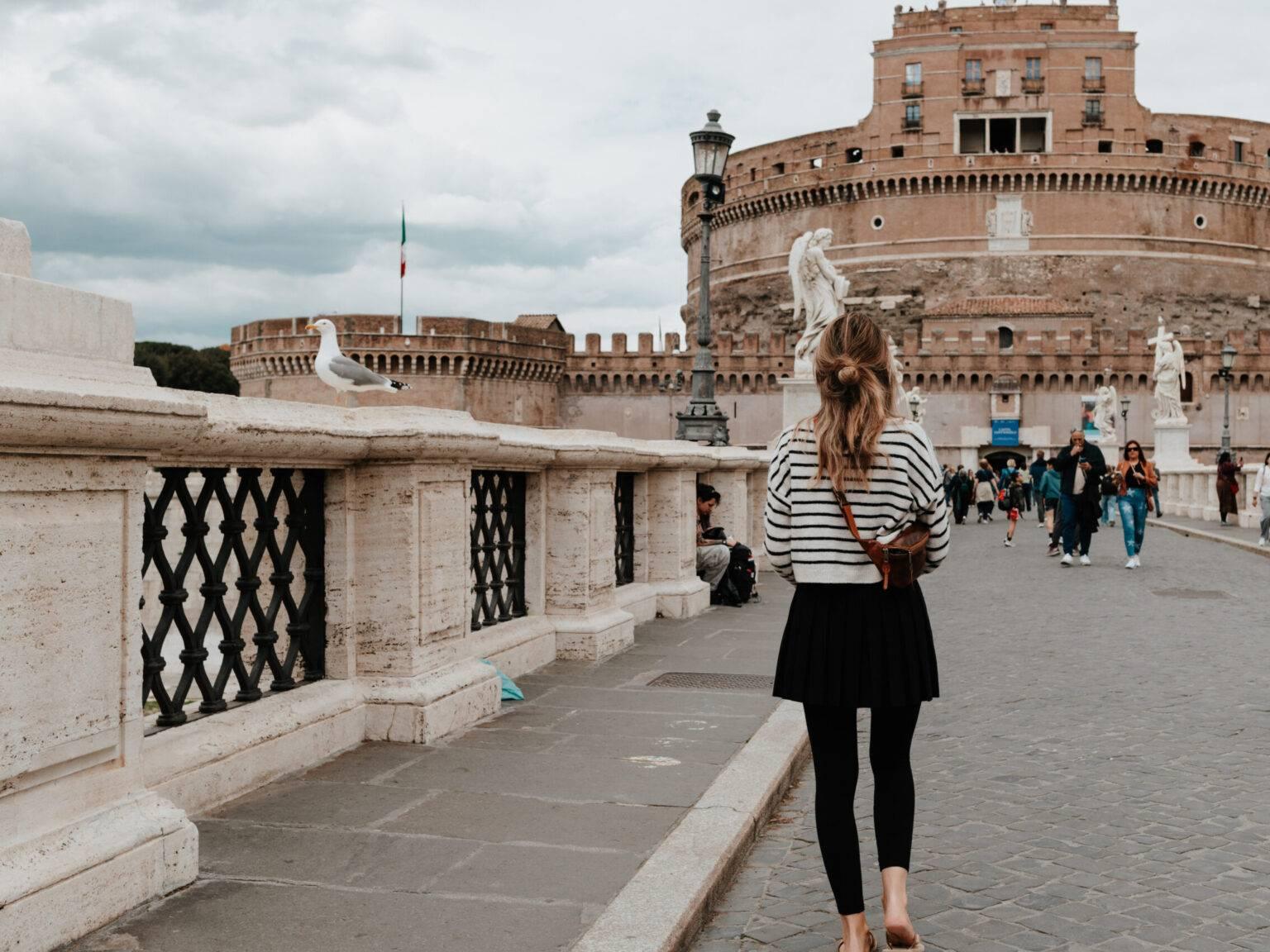 Woman walking across the Angel Bridge in Rome with a view of Castel Sant'Angelo, statues, and a seagull in the foreground.