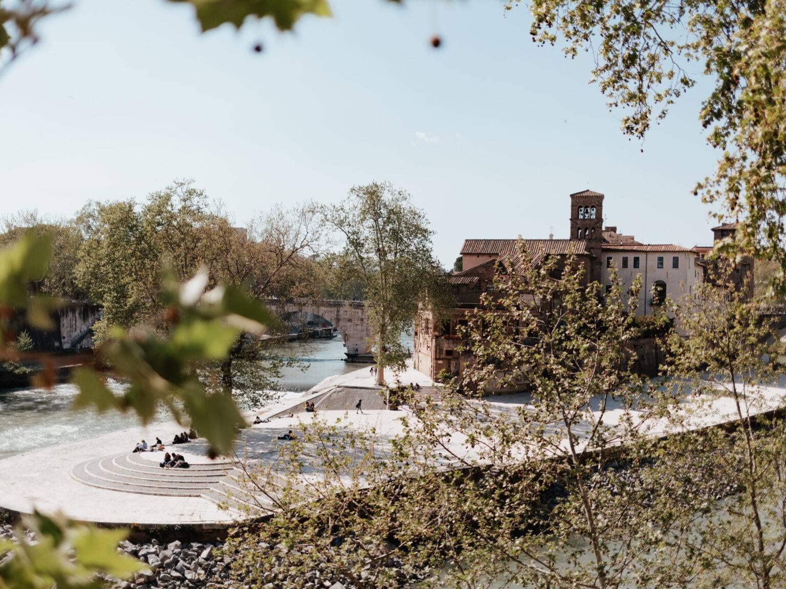Tiber Island in Rome with river, trees, walkers, and historic buildings.