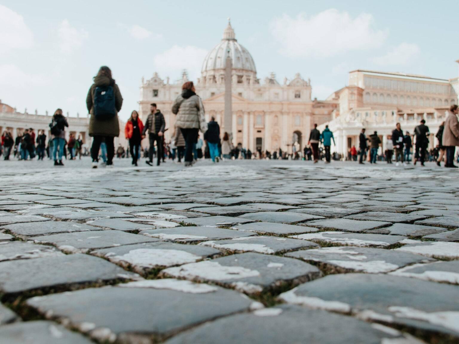 People on St. Peter's Square in Rome with cobblestones and a view of St. Peter's Basilica in the background.