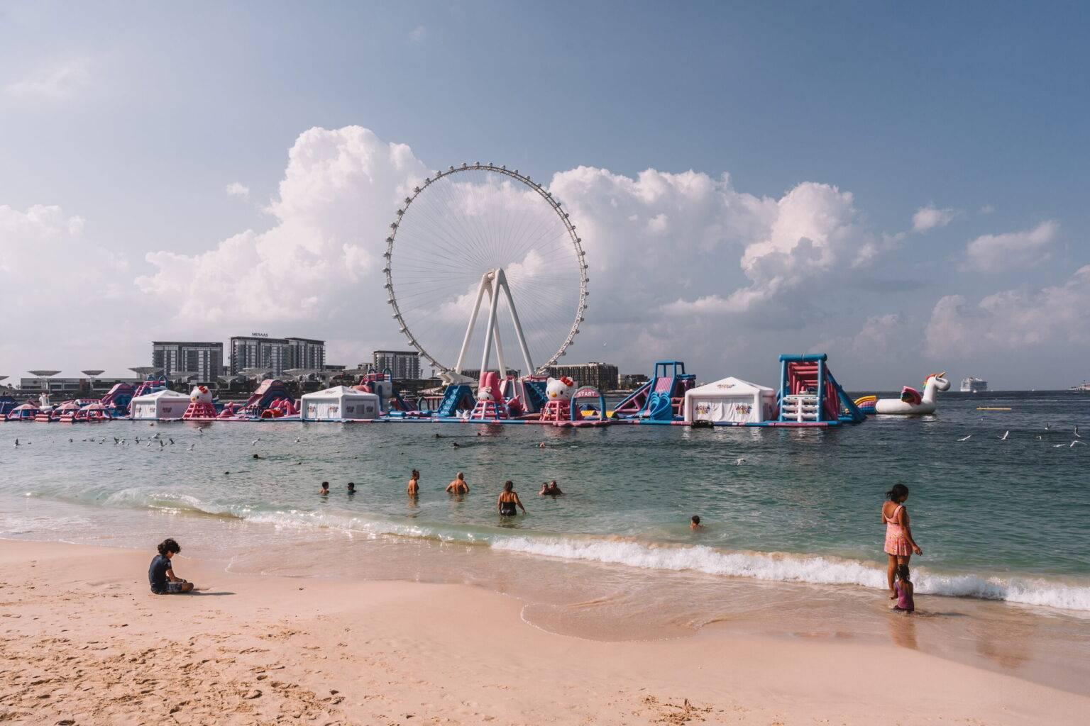 Algunas personas en el mar en la playa JBR en Dubái con toboganes inflables y una noria al fondo.