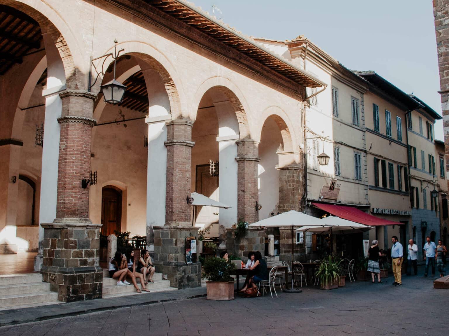 Consejo secreto para los viajeros a la Toscana: el pequeño pueblo de Montalcino - aquí hay arcos donde se sientan algunos jóvenes, al lado un acogedor café con sombrillas en la terraza exterior.