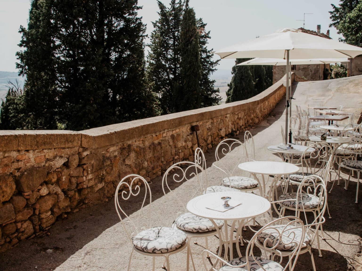 La terraza de un bar invita con sillas blancas de diseño retorcido y sombrillas a disfrutar de la vista del encantador paisaje toscano con sus cipreses.