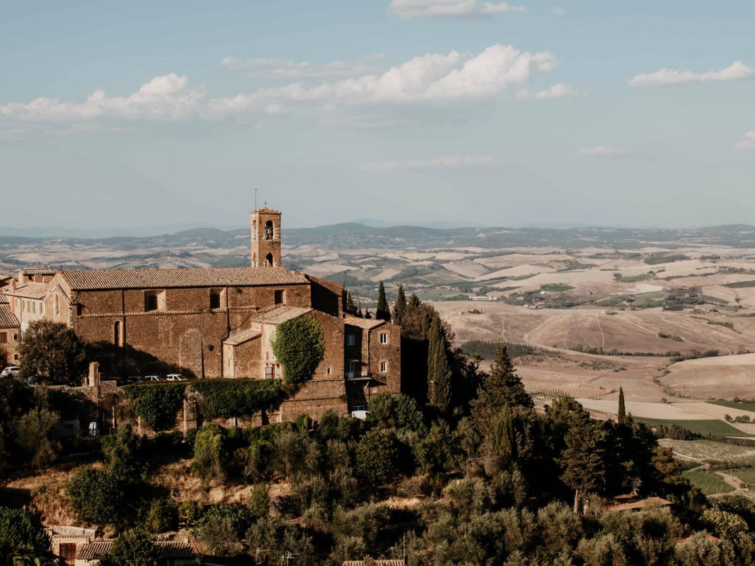 En una elevación se encuentra una fortaleza rodeada de árboles - al fondo el amplio paisaje toscano con sus colinas y cipreses.
