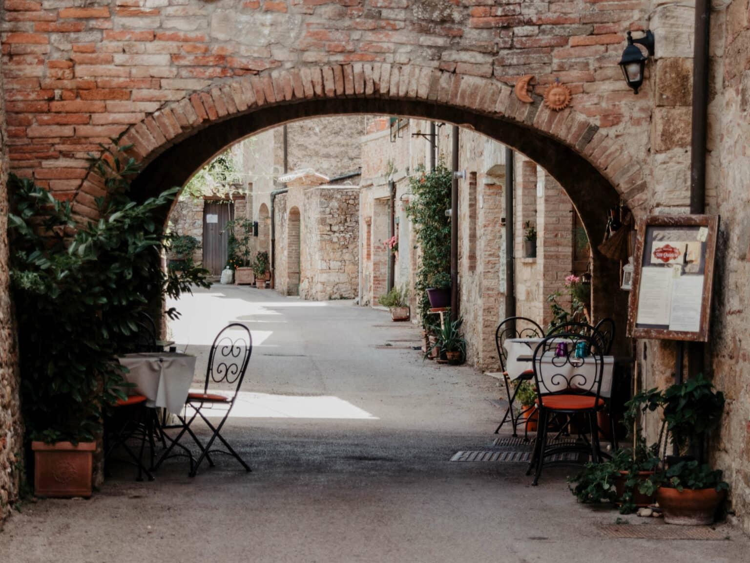 A la sombra del arco de un muro en el pueblo italiano de San Quirico d'Orcia están las mesas y sillas del restaurante.