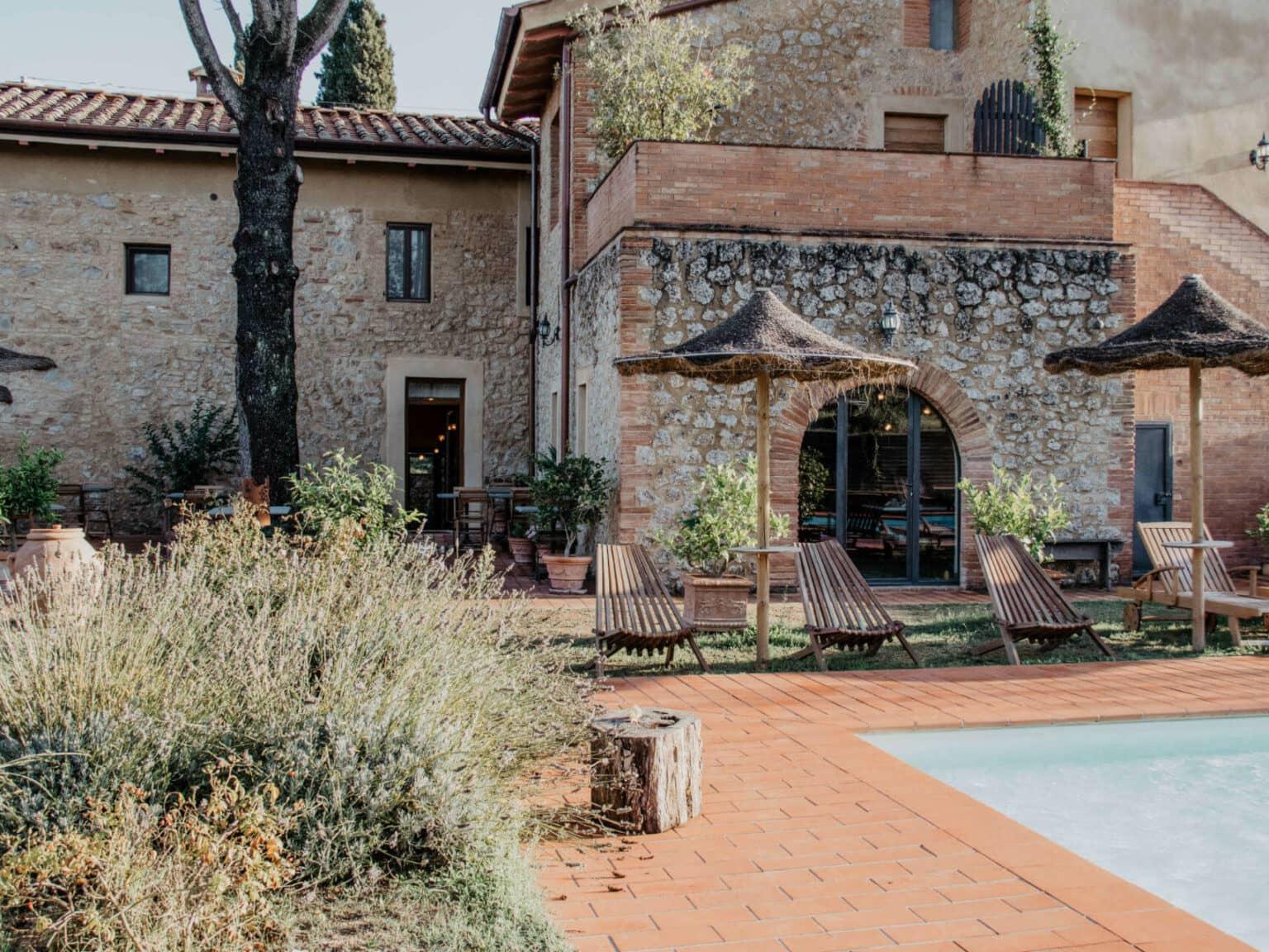 En la piscina de la propiedad Agrivilla i Pini en el corazón de la Toscana, hay tumbonas y sombrillas listas para los vacacionistas, rodeadas de piedras de pavimento rojas.