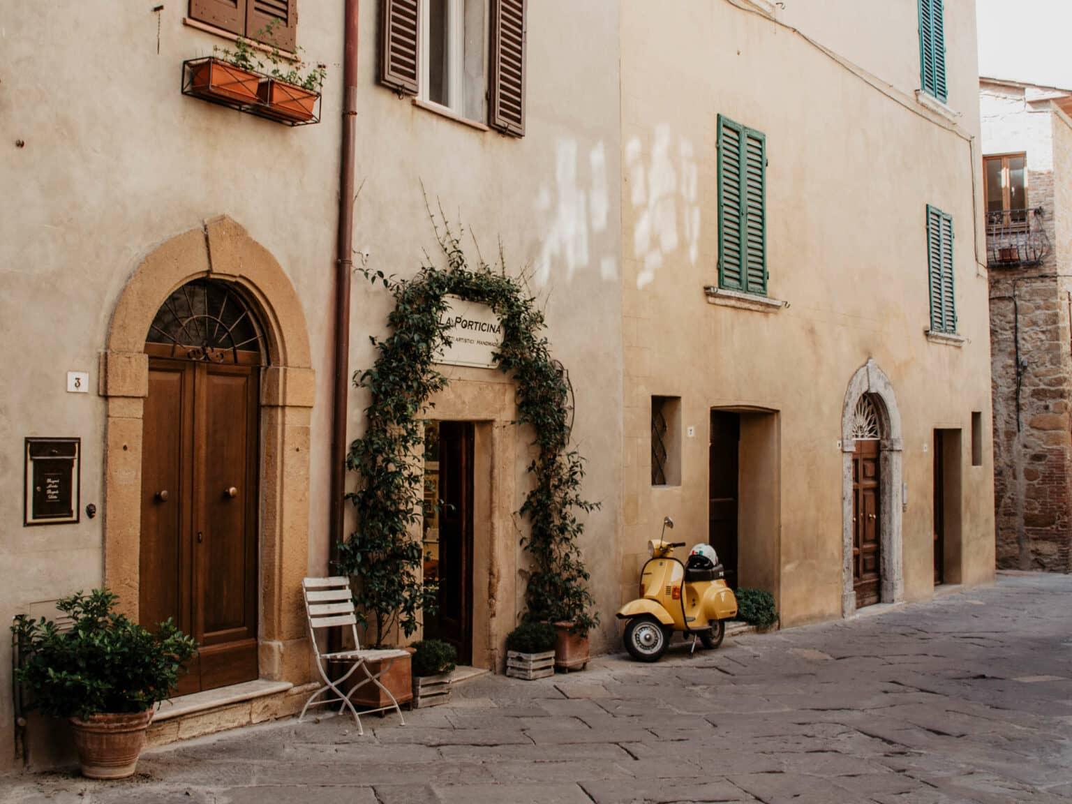 Una Vespa amarilla está apoyada en una casa de arenisca, la pared de la casa está adornada con ventanas de contraventanas verdes, pesadas puertas de madera y una corona de plantas atada a la puerta de entrada.
