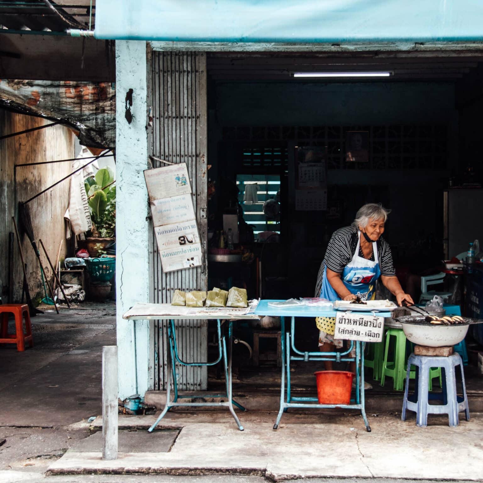 An elderly woman stands behind a street food stall in Bangkok and cooks.