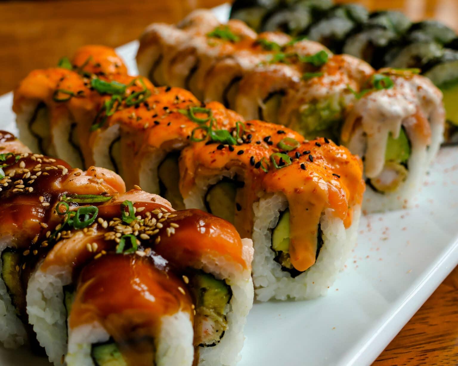 Various sushi rolls topped with sauces on a long white plate.