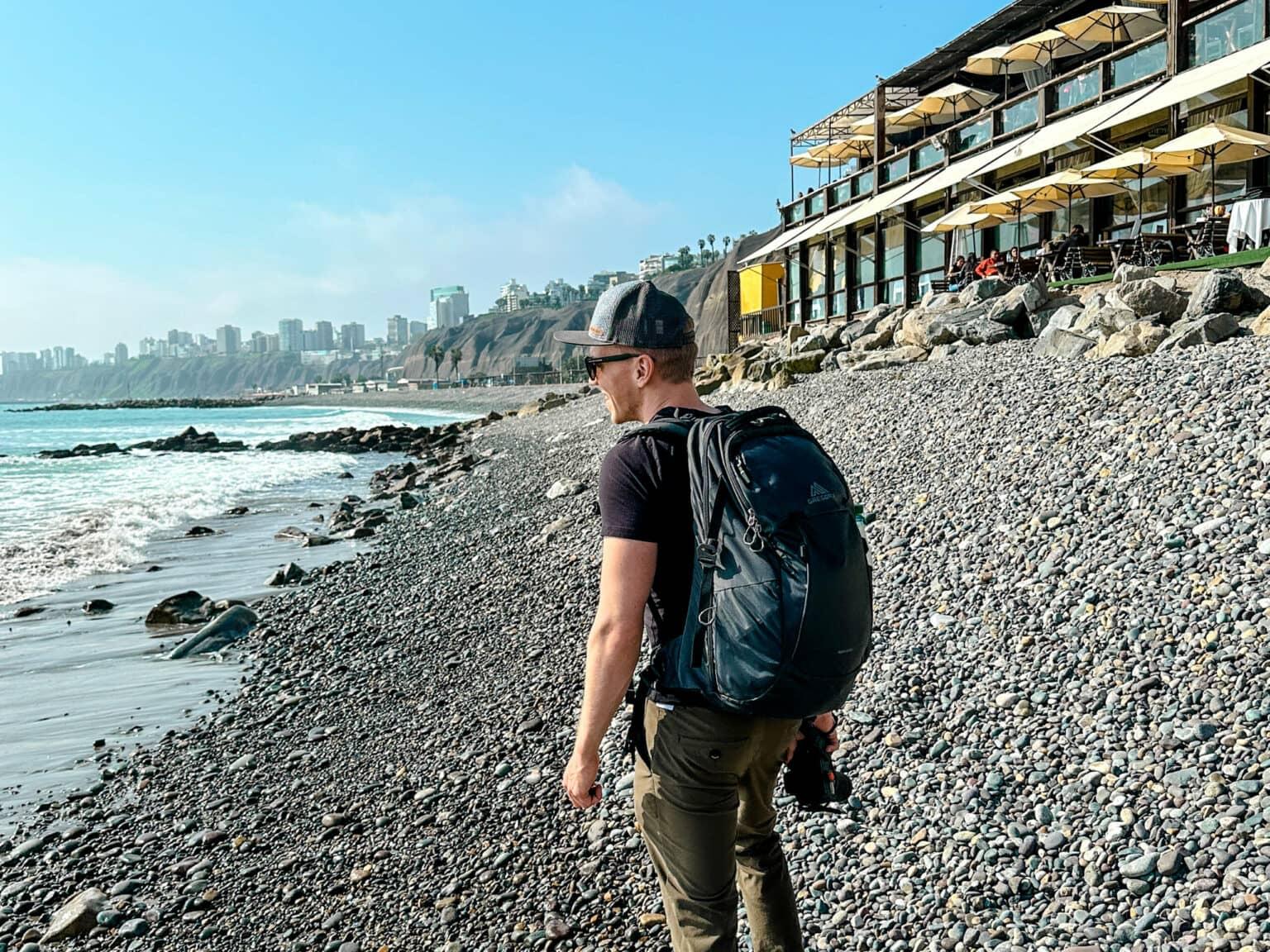 Travel blogger Tom from traveloptimizer stands with a backpack on a pebbly beach in Lima.
