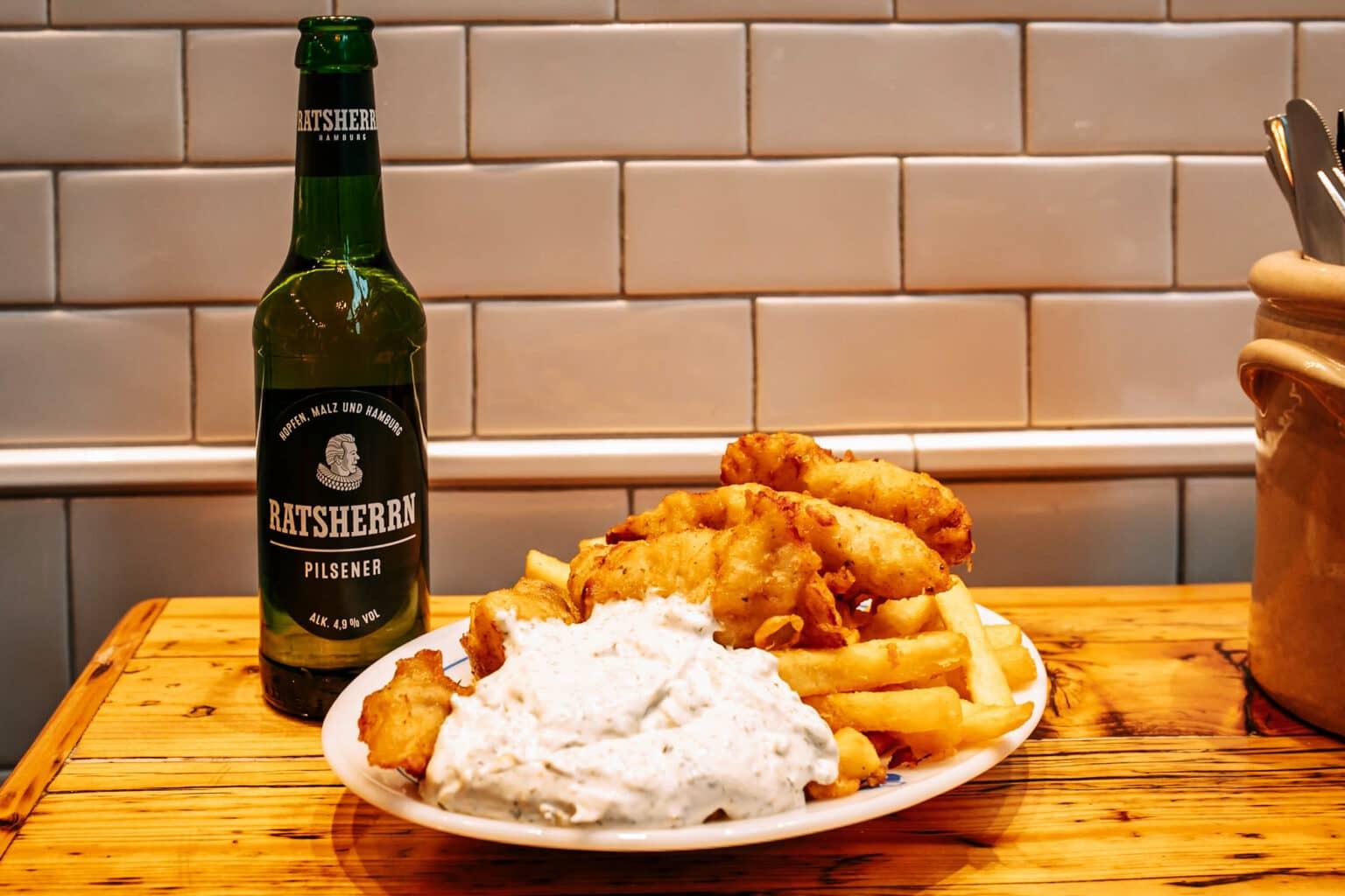 A plate with fish and chips and a bottle of beer on a wooden table at Daniel Wischer by the Hamburg Town Hall.