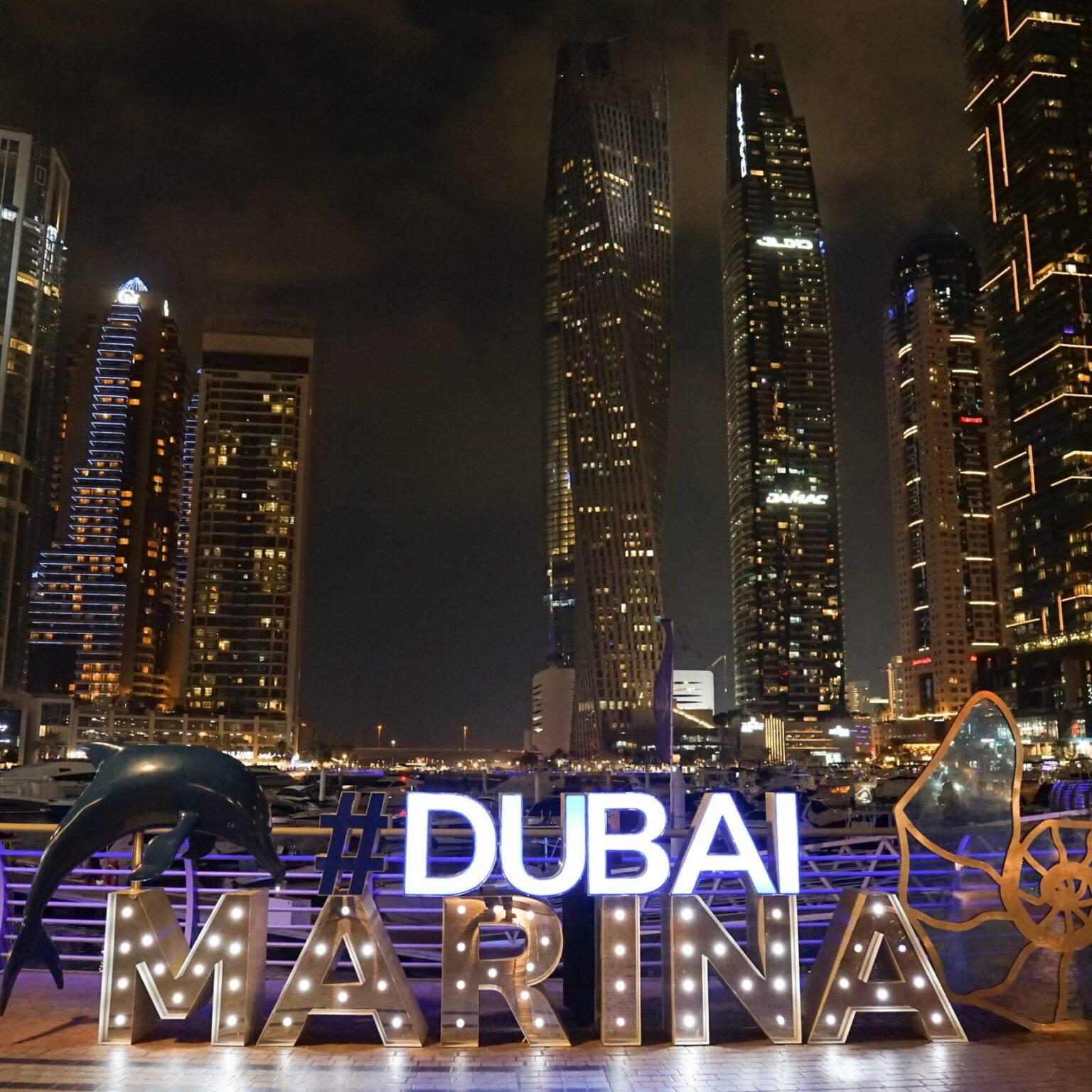Illuminated letters spell out "#DUBAI MARINA". In the background, illuminated skyscrapers at night.