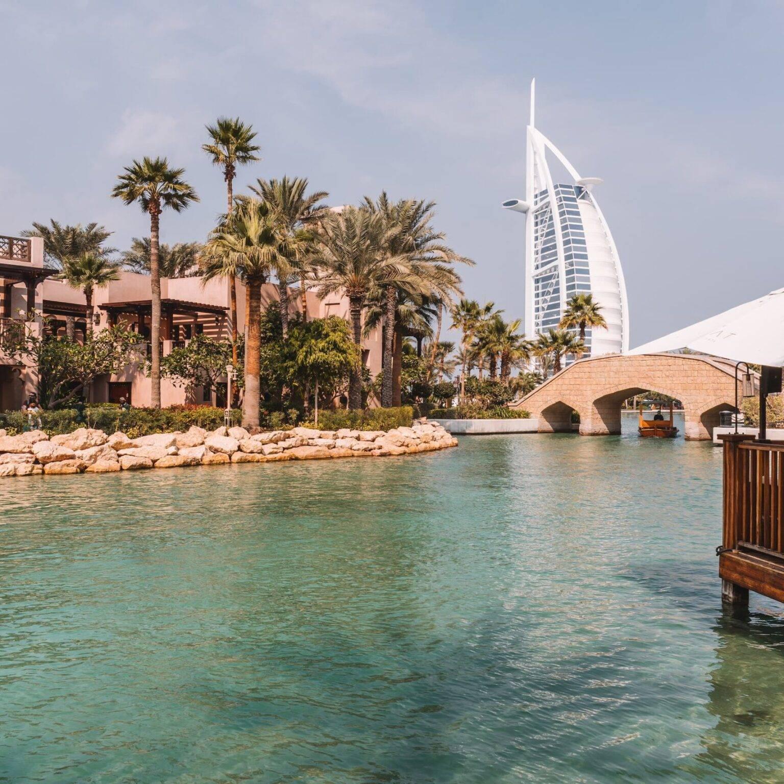 A small river with buildings and palm trees on both sides and the Burj Al Arab Hotel in the background.