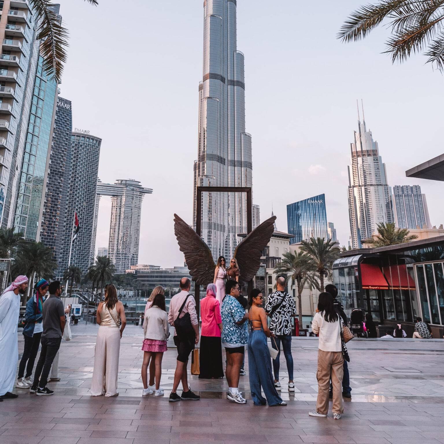 In front of the sculpture "Wings of Mexico", a pair of angel wings made of metal, people are lined up to take photos. The Burj Khalifa stands in the background.