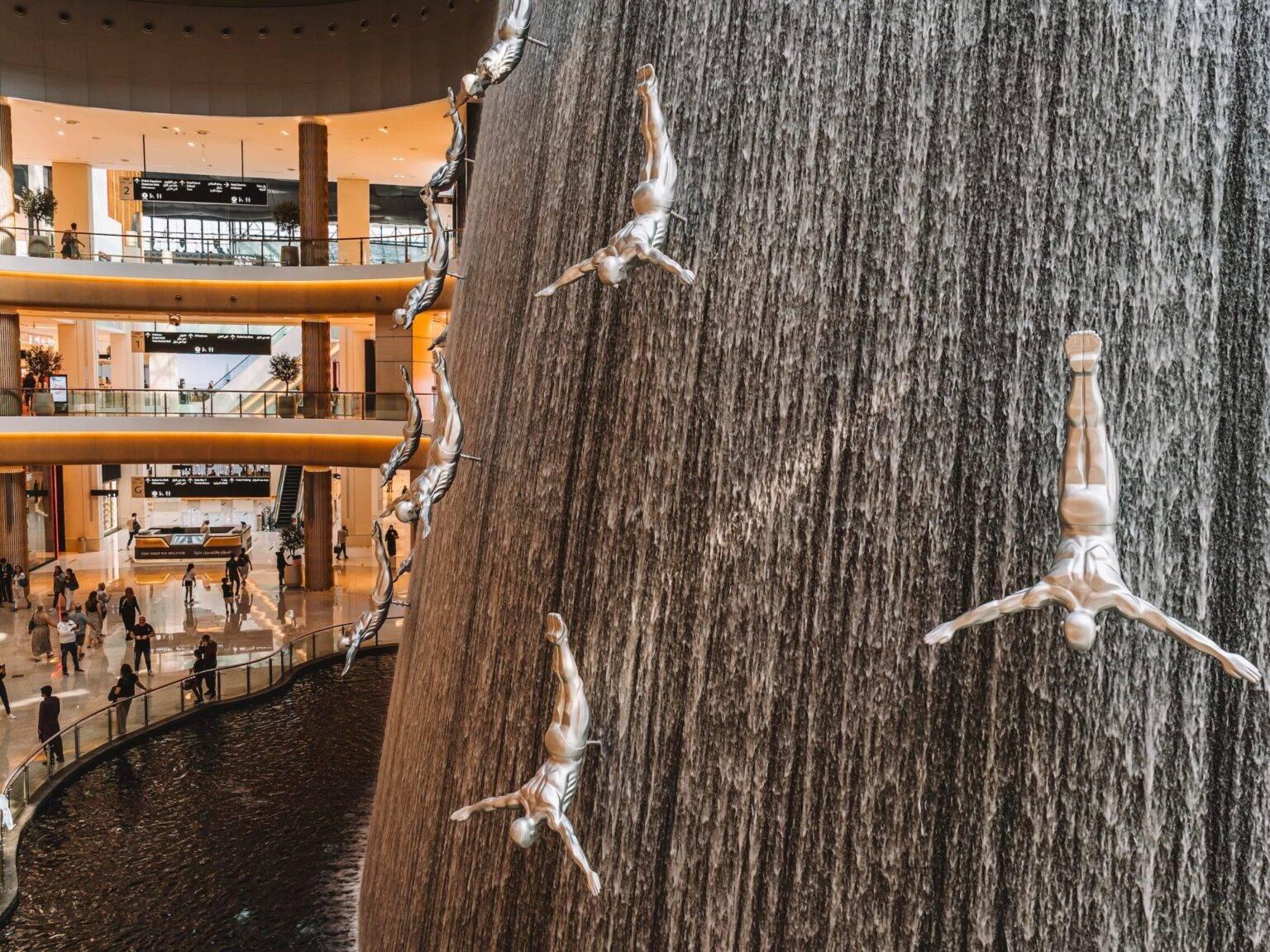 A large artificial waterfall in the Dubai Mall featuring silver male figures diving headfirst and arms outstretched all over the waterfall.