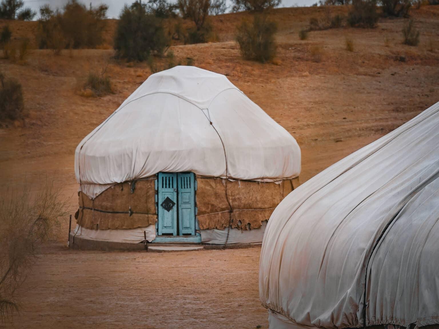 A traditional yurt with a white covering and turquoise wooden door stands in the desert landscape of Uzbekistan