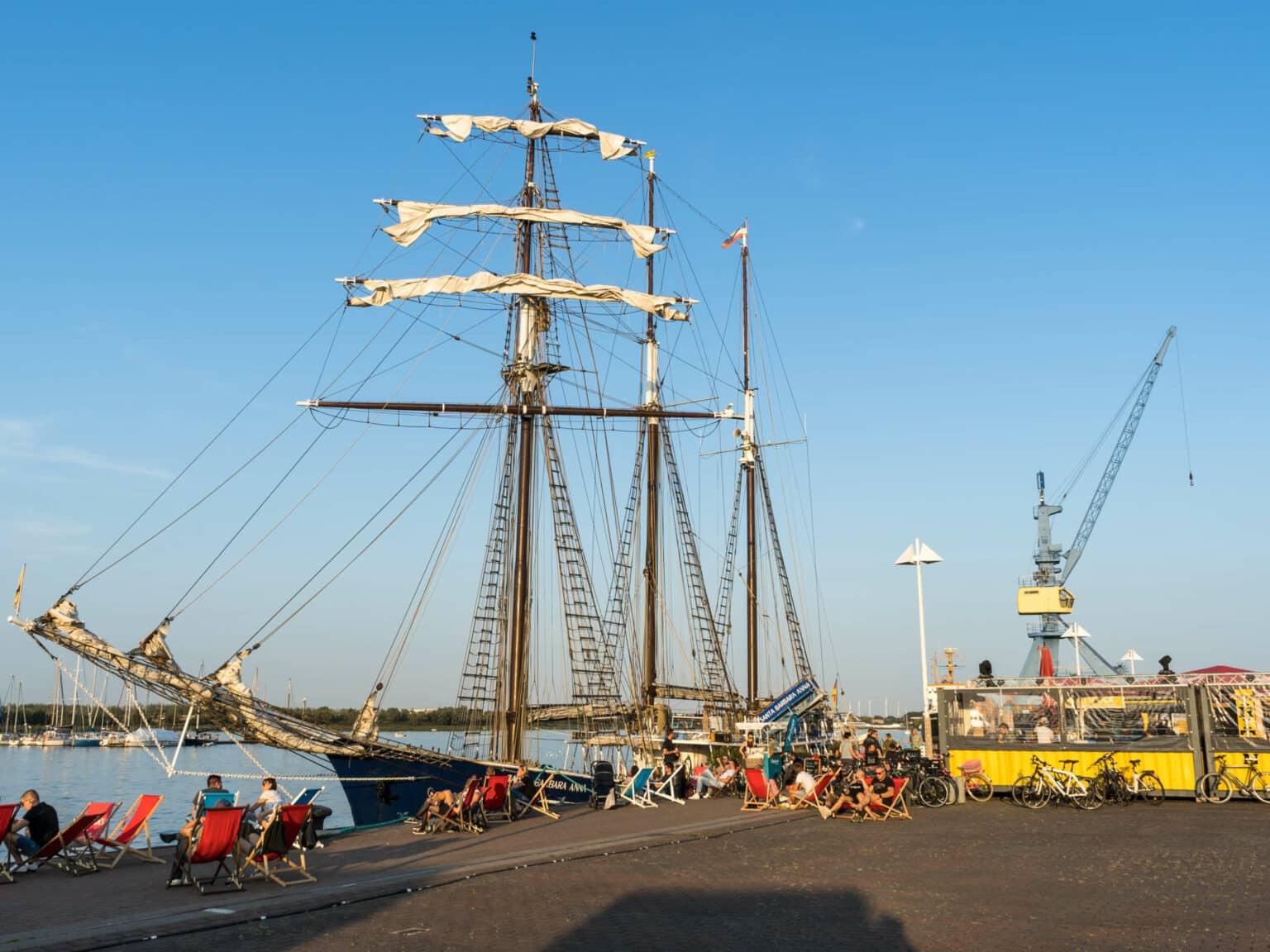 A repurposed ship is docked in Rostock, with people sitting in beach chairs in front, next to a small shelter with leaning bicycles, the sky is bright blue, looking towards a crane.