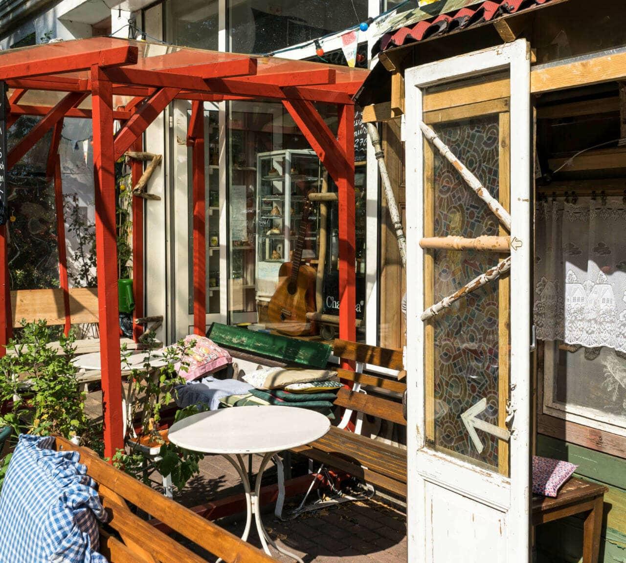 The entrance of a small alternative café in Rostock in the Kröpeliner-Tor-Vorstadt is adorned with a red pergola, wooden benches, cushions, and white tables, an old white wooden door with an arrow points inside the café.