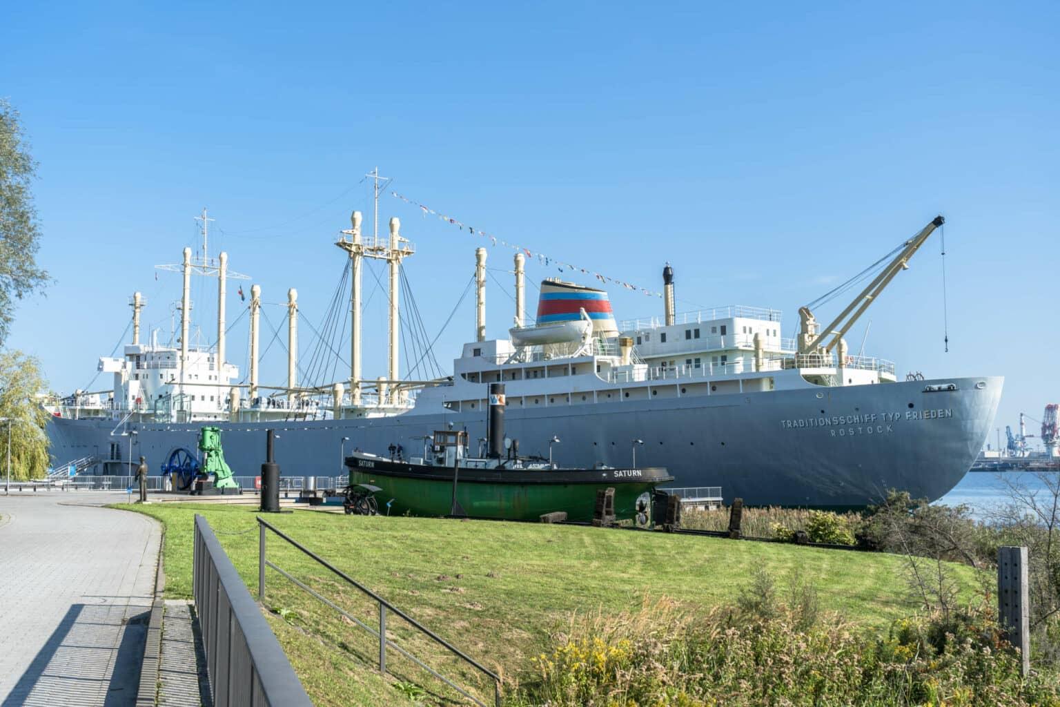 The traditional ship type Frieden is docked by the shore of Rostock, in front of it lies a small green boat, grass, and a cobblestone path, under a bright blue sky.